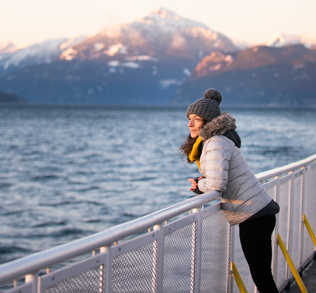A woman looking out over the ocean during a sunset as she leans on a railing. She is wearing winter clothing and there are mountains off in the distance behind her.